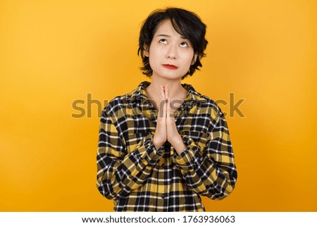 Young beautiful Asian woman with short hair wearing casual plaid shirt over yellow background begging and praying with hands together with hope expression on face very emotional and worried. Asking
