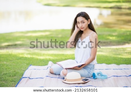 Young beautiful Asian woman sit on mat and hold book to read. Girl picnic by the pond in spring park with leisure lifestyle with copy space for text.