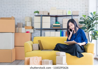 Young And Beautiful Asian Woman Sitting Among Several Boxes And Calling On Yellow Sofa, Working In The House Office. Concept For Home Base Business And Startup Ownership.