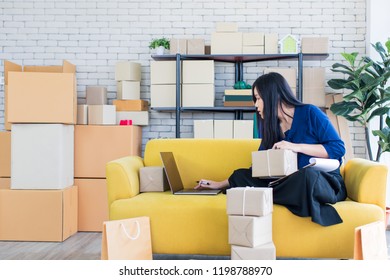 Young And Beautiful Asian Woman Sitting Among Several Boxes And Checking Parcels On The Yellow Sofa, Working In The House Office. Concept For Home Base Business And Startup Ownership.