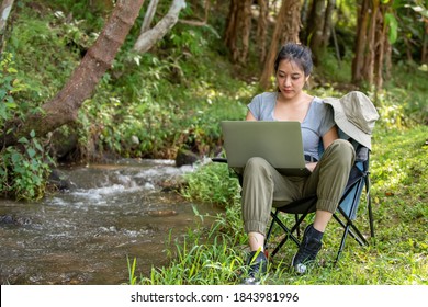 Young Beautiful Asian Woman Sitting On The Chair Using Laptop Computer With Internet For Online Working Outdoor. Pretty Girl Relax And Enjoy Outdoor Lifestyle Camping In Summer Holiday Vacation.