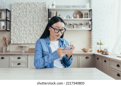 Young Beautiful Asian Woman Sitting In Kitchen At Home And Using Mobile Phone. Scrolls Through The News Feed, Sits On Social Networks, Checks Mail.