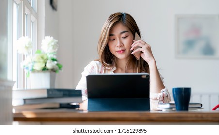 Young Beautiful Asian Woman Using Smartphone And Working With Laptop While Sitting At Office Desk, Working From Home Concept.