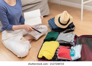 Young Beautiful Asian Woman Using Checklist Prepare Arrange Clothes And Equipment Before Go Travel Oversea After Covid Pandemic. New Normal Travel Lifestyle Concept.