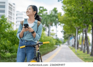 Young beautiful Asian woman is using a smartphone while riding a bicycle through a city park. Lifestyle and environmentally friendly concept. - Powered by Shutterstock