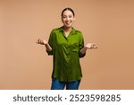 Young, beautiful Asian woman wearing green shirt shrugging with open palms posing in studio isolated on beige background. Advertisement concept