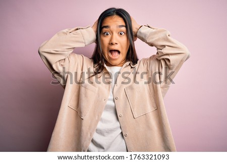 Young beautiful asian woman wearing casual shirt standing over pink background Crazy and scared with hands on head, afraid and surprised of shock with open mouth