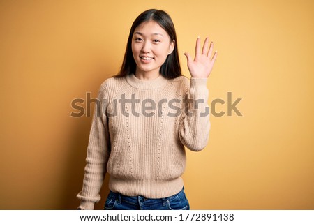 Young beautiful asian woman wearing casual sweater over yellow isolated background Waiving saying hello happy and smiling, friendly welcome gesture