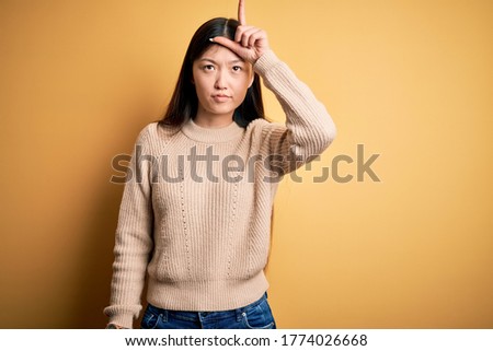 Young beautiful asian woman wearing casual sweater over yellow isolated background making fun of people with fingers on forehead doing loser gesture mocking and insulting.