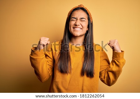 Young beautiful asian woman wearing casual sweater and diadem over yellow background very happy and excited doing winner gesture with arms raised, smiling and screaming for success. Celebration