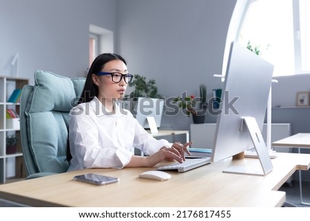 A young beautiful Asian woman is working at a computer in the office. Businesswoman, director, freelancer, manager, sitting in a white shirt and glasses at the desk.