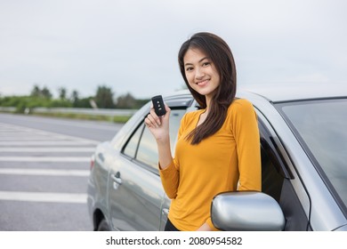 Young Beautiful Asian Women Buying New Car. She Was Standing In Near Car On The Roadside. Hand Showing Car Key. Smiling Female Driving Vehicle On The Road On A Bright Day