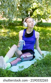 Young Beautiful Athletic Woman With Bionic Prosthetic Leg Doing Sport Exercises In The Park Outdoors In Summer. Concept Of Active Lifestyle Of Disabled People. Listening To Music, Drinking Water