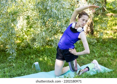 Young Beautiful Athletic Woman With Bionic Prosthetic Leg Doing Sport Exercises In The Park Outdoors In Summer. Concept Of Active Lifestyle Of Disabled People