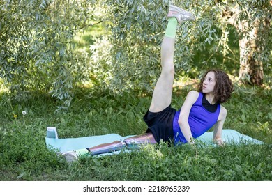 Young Beautiful Athletic Woman With Bionic Prosthetic Leg Doing Sport Exercises In The Park Outdoors In Summer. Concept Of Active Lifestyle Of Disabled People