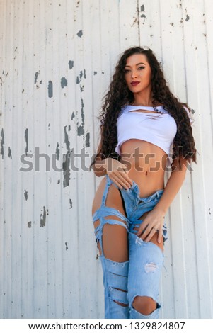 Young beautiful athletic woman in a short vest and blue ripped jeans posing against a white lighthouse on a warm summer day near the sea. Girl walks near the sea