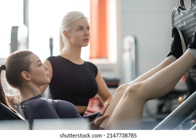 A Young Beautiful Athletic Woman Trains In The Gym With The Help Of A Female Coach. Leg Press On The Training Apparatus. Gym.