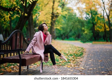 Young beautiful ballerina in fair coat and pointe shoes sit on the bench, rest outdoors in autumn park. - Powered by Shutterstock