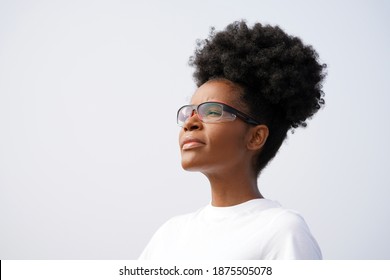 Young beautiful black scientist standing against sky outside with protective glasses, a white shirt, and a high puff afro ponytail hairstyle, looking up and to the left                               
