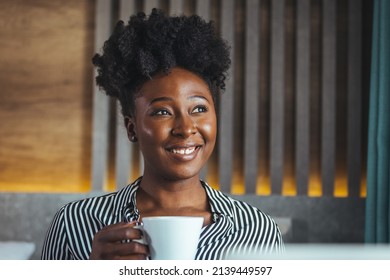 Young beautiful black woman with curly hair woman in a hotel with a white striped shirt drinking coffee while sitting on the bed in the morning and drinking coffee.