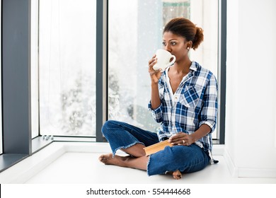 young beautiful black woman lying on the floor and drinking coffee or tea and reading a book next to big windows with natural light - Powered by Shutterstock