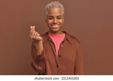 Young Beautiful Blond African American Woman Smile And Makes Korean Love Sign, Shapes Heart With Her Fingers Expresses Her Love And Sympathy, Stand Over Brown Background. Hand Gesture Concept.