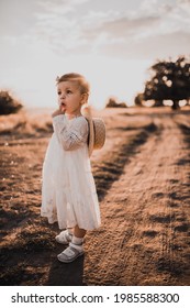 young beautiful blond girl baby straw hat white dress bow on head background dry field meadow middle steppe outdoors. little toddler child walks alone. Romantic rural image pure nature Summer sunset