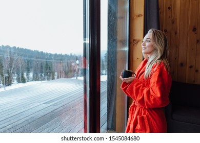 Young beautiful blond woman dressed in red bathrobe standing near big window holding cup of coffee and watching first snow falling. - Powered by Shutterstock