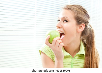 Young beautiful blond woman eat green apple with venetian blind window background