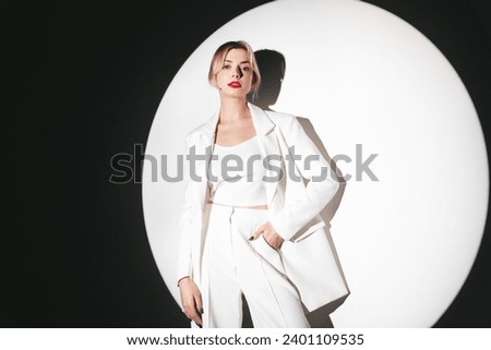 Young beautiful blond woman wearing nice trendy white suit and jacket. Fashion model posing in studio. Fashionable female posing near white wall in a circle light. Cheerful and happy. With red lips