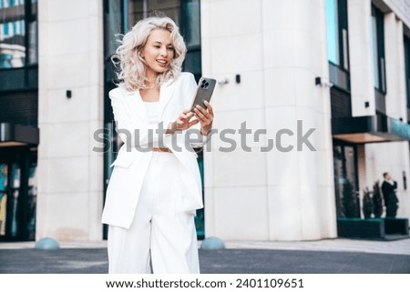 Young beautiful blond woman wearing nice trendy white suit jacket. Smiling model posing in street. Fashionable female outdoors. Cheerful and happy. Holds smartphone, uses phone apps, looks at screen