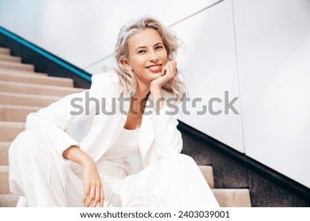 Young beautiful blond woman wearing nice trendy white suit jacket. Smiling model posing in the street at sunny day. Fashionable female outdoors. Cheerful and happy. Sits at the stairs 