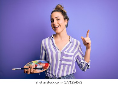 Young beautiful blonde artist woman painting using brush and palette over purple background surprised with an idea or question pointing finger with happy face, number one