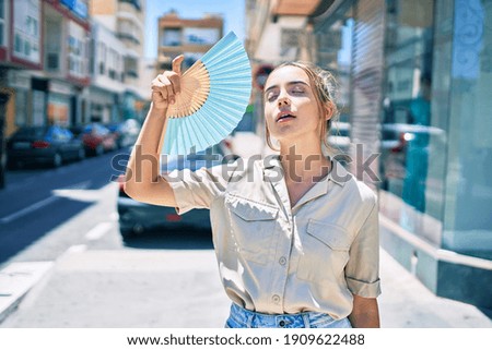 Young beautiful blonde caucasian woman smiling happy outdoors on a sunny day using handfan for hot weather