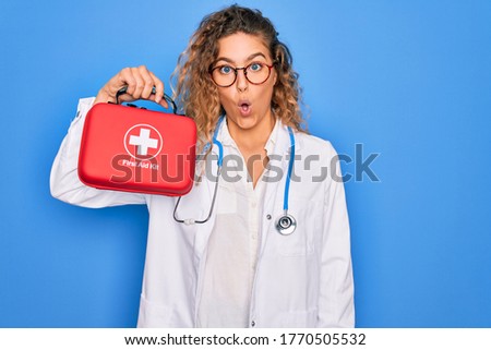 Young beautiful blonde doctor woman with blue eyes wearing stethoscope holding first aid kit scared in shock with a surprise face, afraid and excited with fear expression