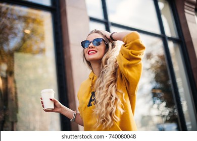young beautiful blonde drinking coffee walking around the city.mustard sweetshot.,urban backpack , bright red lips Posing against the window of the boutique Model looking aside and smile
