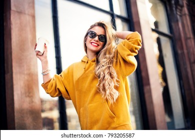 young beautiful blonde drinking coffee walking around the city.mustard sweetshot.,urban backpack , bright red lips Posing against the window of the boutique.