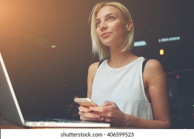 Young beautiful blonde female holding mobile phone while sitting with portable net-book in coffee shop interior, charming dreamy woman using cell telephone and laptop computer during rest in cafe 