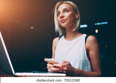 Young beautiful blonde female holding mobile phone while sitting with portable net-book in coffee shop interior, charming dreamy woman using cell telephone and laptop computer during rest in cafe