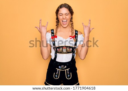 Young beautiful blonde german woman with blue eyes wearing traditional octoberfest dress shouting with crazy expression doing rock symbol with hands up. Music star. Heavy music concept.