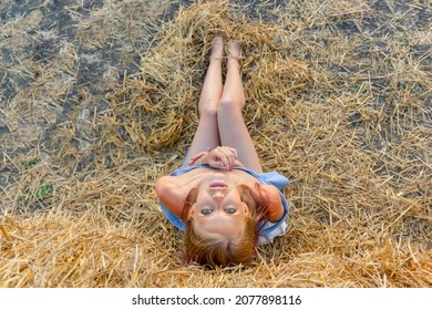 A young beautiful blonde girl with blue eyes and snow-white skin sits on the hay on the ground and her head thrown back looks relaxed up. Summer sunny day