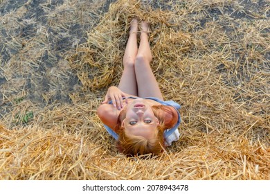 A young beautiful blonde girl with blue eyes and snow-white skin sits on the hay on the ground and her head thrown back looks relaxed up. Summer sunny day