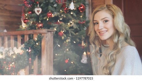 Young beautiful blonde girl with curly hair smiling and looking at Christmas tree. Portrait of woman in warm sweater. New year concept shot on bright background with lens flares. 