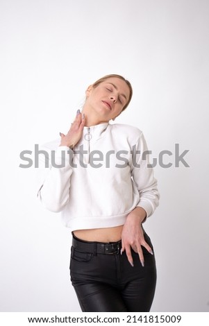 Young beautiful blonde girl on a white background in a white sweatshirt and black pants, hand around her neck