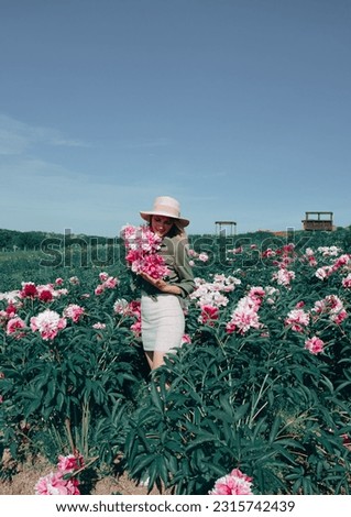 Young beautiful blonde girl with pink and white peonies in her hands, sunny summer day, cropped image, field path background, girl in dress in field.Beautiful young girl among peonies, peony field