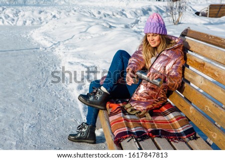 Young beautiful blonde girl sits on a bench in winter park in with thermos drinking hot tea