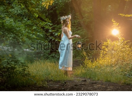 a young beautiful blonde hair  girl is standing in the forest in a white long summer dress barefoot, she has a wreath of daisies on her head and an old kerosene lamp in her hands
