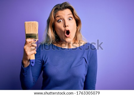 Young beautiful blonde painter woman painting using paint bush over purple background scared in shock with a surprise face, afraid and excited with fear expression