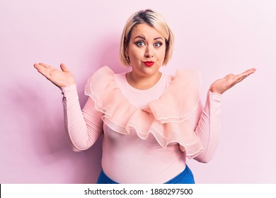 Young Beautiful Blonde Plus Size Woman Wearing Casual Sweater Over Isolated Pink Background Clueless And Confused With Open Arms, No Idea And Doubtful Face.