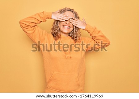 Young beautiful blonde sporty woman wearing casual sweatshirt over yellow background covering eyes with hands smiling cheerful and funny. Blind concept.